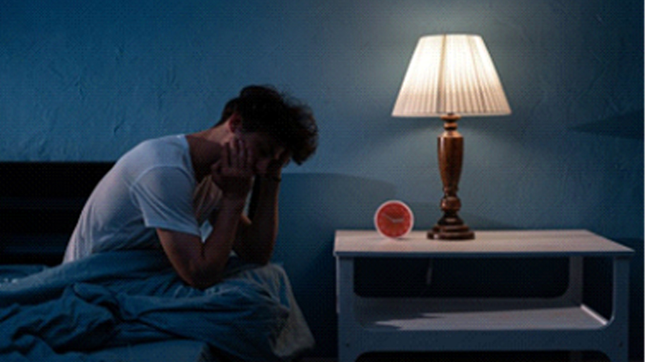 Man sitting up in bed with the lamp on and is unable to sleep